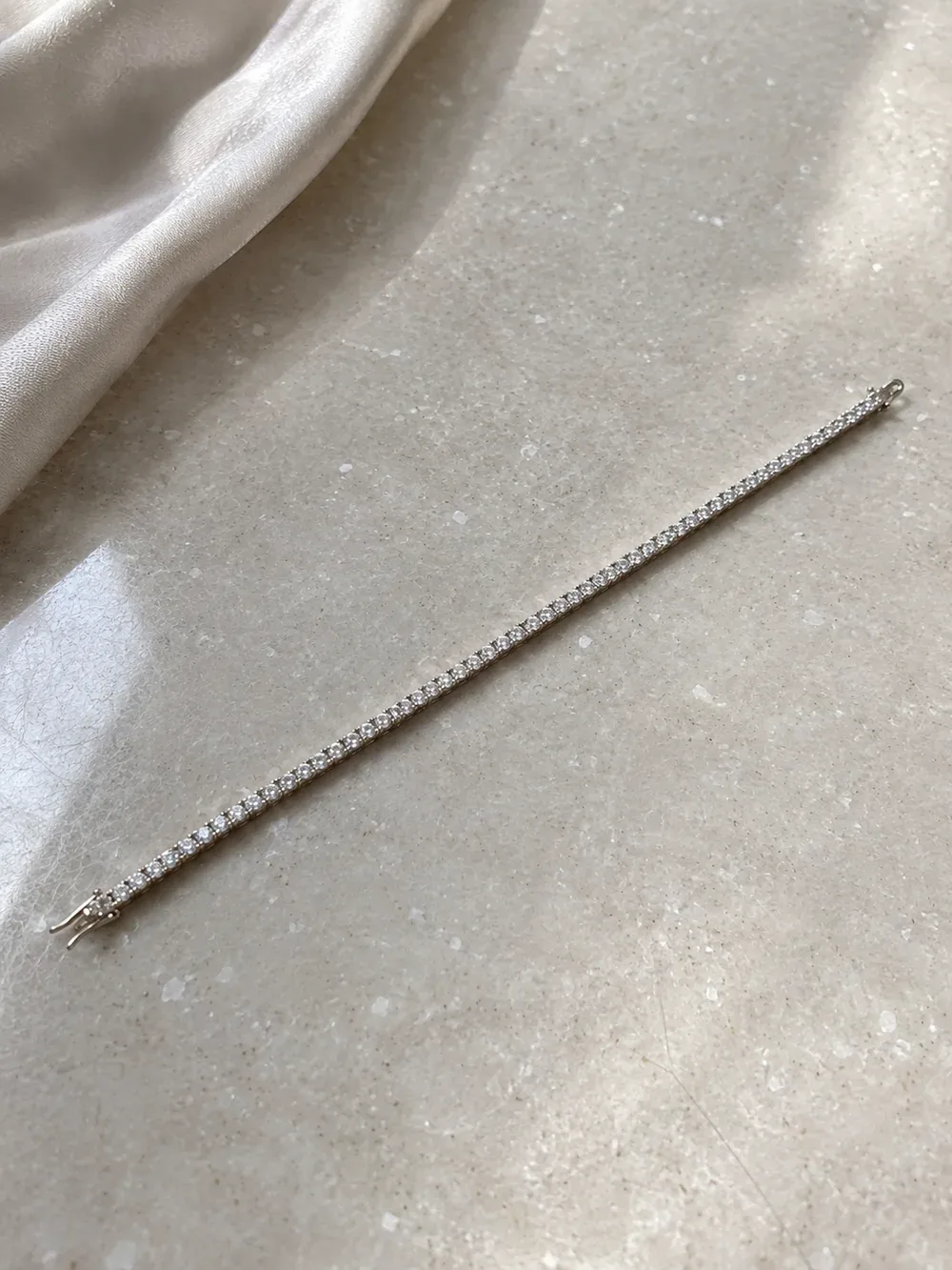 A white gold diamond tennis bracelet laid on cream marble beside silk